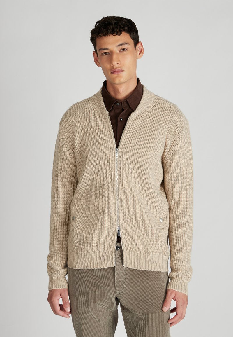 Beige geribde cardigan met een rits aan de voorkant, met lange mouwen en twee zijzakken. Draagt over een donkerbruine shirt, tegen een neutrale achtergrond.