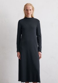 Czarna sukienka z długim rękawem, wykonana z miękkiego materiału, z wysokim dekoltem i falbankowym wykończeniem. Prosta sylwetka o luźnym kroju, bez widocznych wzorów.