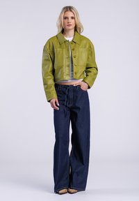 Przycięta zielona skórzana kurtka z dużymi guzikami, w połączeniu z wysokim stanem ciemnoniebieskimi spodniami o szerokich nogawkach i beżowym obuwiem.