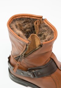 Botte en cuir marron avec fermeture éclair ouverte, révélant une doublure intérieure douce et pelucheuse ainsi qu'une sangle marron foncé ornée de clous en métal autour de la cheville.