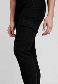 Pantalones cargo de mezclilla negra con un bolsillo lateral, diseño ajustado y textura suave; detalle de cremallera visible en la cadera.