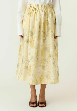 Donna che indossa una lunga gonna floreale giallo pastello con vita elastica, top bianco e tacchi aperti marroni.