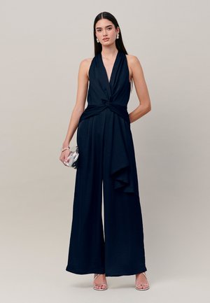 Vrouw met lang donker haar, die een mouwloze marineblauwe jumpsuit met wijde pijpen en een zilveren clutch draagt, staande tegen een effen lichte achtergrond.
