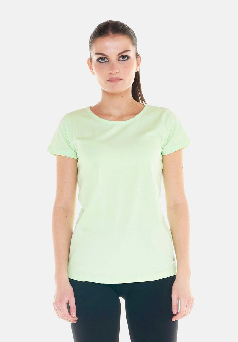 Jeune femme aux cheveux foncés en queue de cheval, portant un t-shirt vert clair à manches courtes et un pantalon foncé, debout devant un fond blanc.