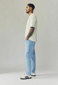T-shirt beige chiaro, jeans dritti blu chiaro e sneakers bianche con strisce nere. Il soggetto ha capelli corti e mossi e tatuaggi visibili.