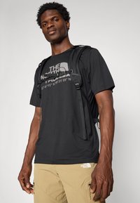 T-shirt noir à manches courtes avec un design de logo réfléchissant, associé à un pantalon cargo beige avec des zips et un logo de marque blanc sur la jambe.