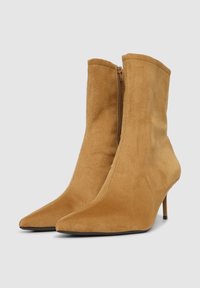 Tannfarbene Wildleder-Stiefeletten mit spitzen Zehen, einem seitlichen Reißverschluss und einem Stiletto-Absatz. Die glatte Textur und die taillierte Silhouette unterstreichen das Design.