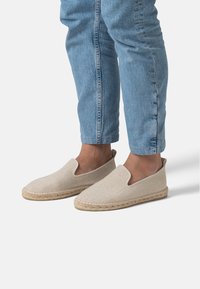 Scarpe espadrillas beige senza lacci con texture intrecciata, pannelli laterali elastici e suola in juta, indossate con jeans azzurri arrotolati.