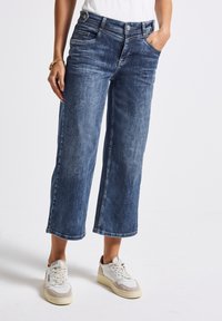 Breda ben mörkblå denimjeans med en urtvättad finish, utrustad med framfickor och denimstickning. Matchas med vita och beige sneakers.