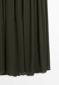Dunkelgrünes, plissiertes Kleid aus leichtem Stoff mit einem Kordelzug in der Taille und einem fließenden, weichen Textursaum.