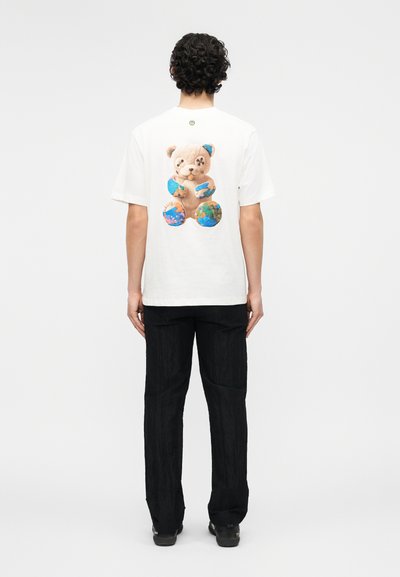 Άνδρας όρθιος με την πλάτη να φαίνεται, φοράει λευκό μπλουζάκι με πολύχρωμο σχέδιο αρκουδάκι στην πλάτη, μαύρο παντελόνι και μαύρα παπούτσια.