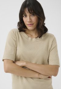 Beige krátký rukáv top s kulatým výstřihem, měkkou texturou a jemným žebrováním, modelovaný osobou se zkříženýma rukama.