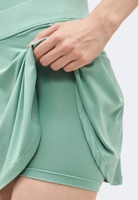 Χέρι σηκώνει φούστα σε μέντα χρώμα για να αποκαλύψει εφαρμοστά σορτς από κάτω, τονίζοντας τα στρωμένα υφάσματα αθλητικής ένδυσης και τις λεπτομέρειες των ραφών.