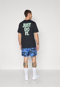 T-shirt in cotone nero con la scritta "JUST DO IT." in verde sul retro, abbinato a pantaloni corti blu a fantasia, calze bianche e sneakers.