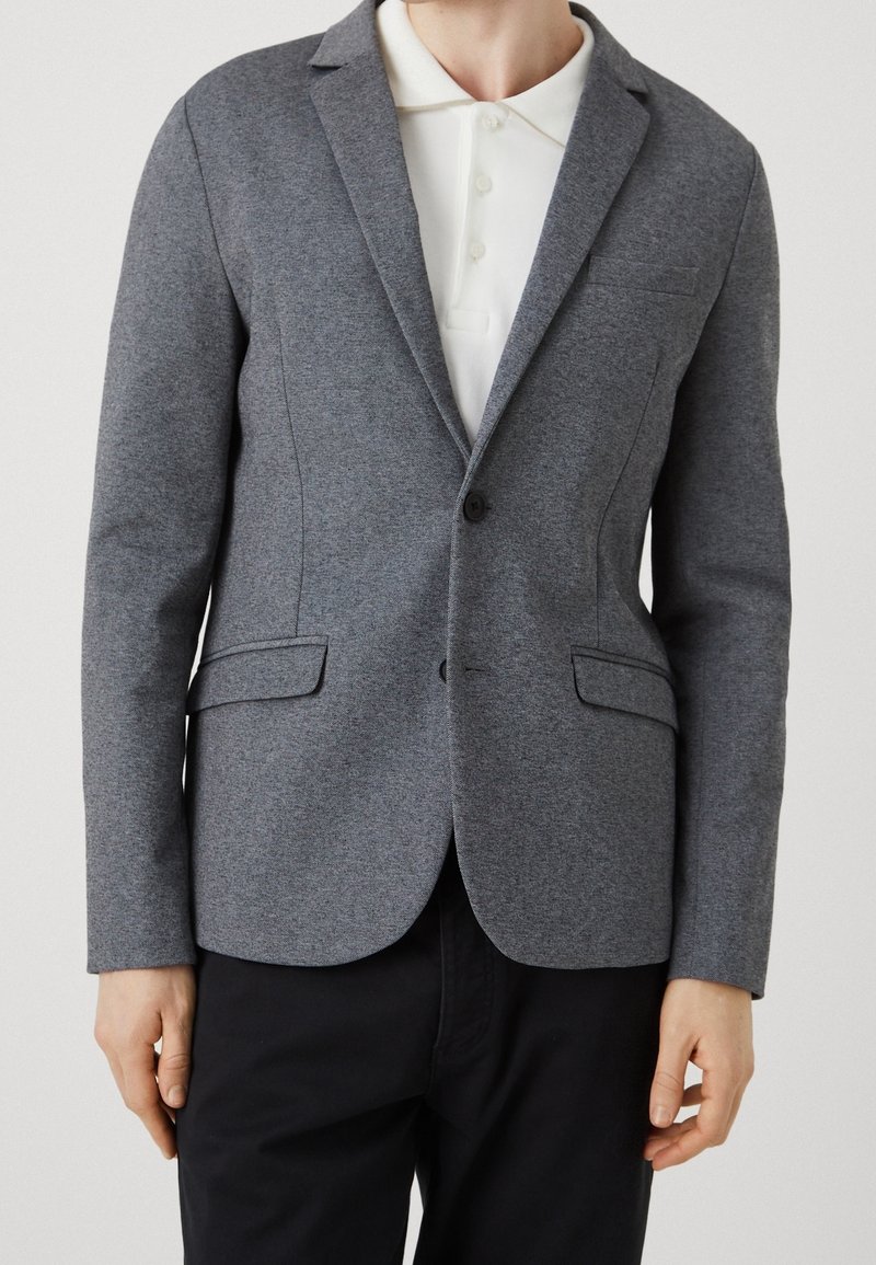 Blazer grigio realizzato in tessuto testurizzato, con revers a punta, chiusura con un solo bottone e tasche laterali, indossato sopra una camicia bianca con colletto.