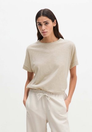 Vrouw met lang donker haar die een beige T-shirt met korte mouwen en lichtbeige broek met trekkoord draagt, staand met haar handen in haar zakken.