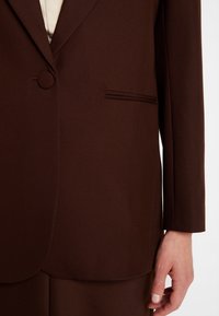 Hnědý přizpůsobený blazer vyrobený z texturované látky. Mezi pozoruhodné rysy patří kulaté knoflíky, jedno náprsní kapsa a čisté linie.