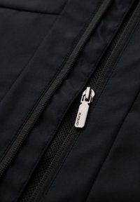 Nahaufnahme eines schwarzen Jackenreißverschlusses mit einem silbernen Metall-Zippergriff, auf dem das Wort "ombre" eingraviert ist, auf Stoff- und Meshfutter.