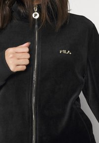 Žena v černé sametové bundě s zlatým logem FILA, držící táhlo zipu blízko límce.