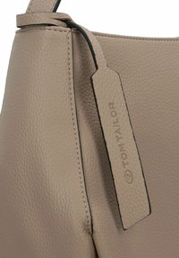Beige Handtasche mit strukturiertem Material und schwarzem Akzentgurt. Verfügt über ein Etikett mit eingeprägtem "TOM TAILOR"-Logo. Glattes, strukturiertes Design.