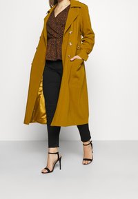 Trench coat giallo senape con bottoni doppi, tasche laterali e foderato in satin; abbinato a una camicetta a fantasia e pantaloni neri.