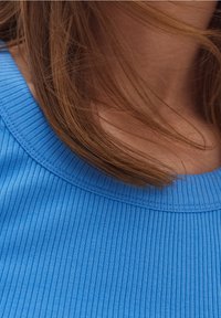 Ribbed modrá blůza s kulatým výstřihem, která ukazuje hladkou texturu a krátké vlasy jemně rámující límec.