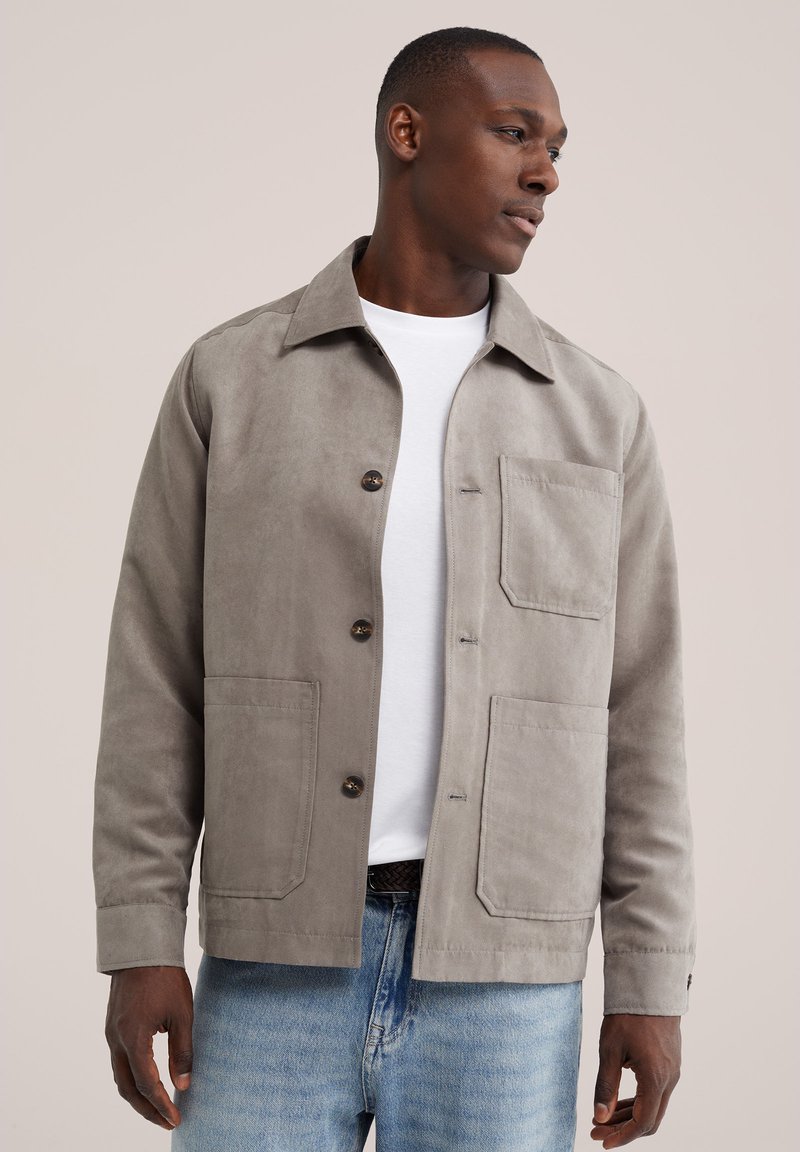 Homme portant une veste boutonnée gris clair par-dessus un t-shirt blanc et un jean bleu, regardant vers sa droite sur un fond clair uni.