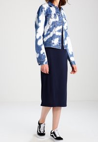 Jeansowa kurtka z niebiesko-białym wzorem tie-dye, noszona na granatowej sukience midi. W połączeniu z czarno-białymi trampkami, stojąca na jednolitym tle.
