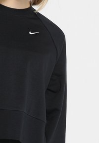 Czarna bluza Nike z długim rękawem noszona przez osobę, z białym logo Nike swoosh na górnej części klatki piersiowej, blisko ramienia.