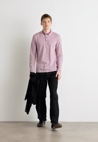 Polo majica s dugim rukavima, svijetlo ružičaste boje, s dvije tipke i bijelim ovratnikom, u kombinaciji s crnim hlačama i crnom jaknom prebačenom preko jednog ramena.