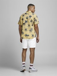 Korte mouwen gele shirt met een marineblauw palmboom patroon, gecombineerd met witte shorts, witte sokken met rode en zwarte strepen, en grijze sneakers.