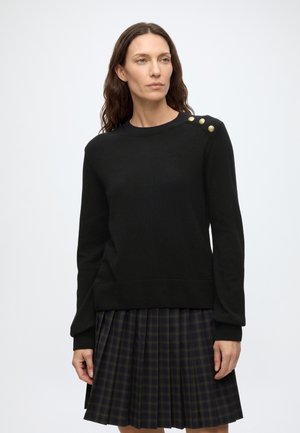 Frau mit langen braunen Haaren, die einen schwarzen Pullover mit drei goldenen Knöpfen auf der Schulter und einen dunkel karierten plissee Rock trägt.