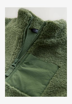 Veste en polaire vert olive avec col montant, fermeture éclair et surface texturée ; présente une poche carrée en tissu lisse.