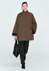 Cappotto overesize marrone con colletto alto, bottoni neri e polsini a contrasto neri. Abbinato a pantaloni a righe e a una borsa nera.