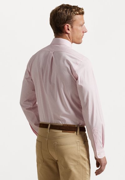 Camisa de dress de rayas rosa claro con mangas largas, un pliegue en la parte de atrás y puños abotonados, combinada con pantalones beige que tienen un cinturón texturizado.