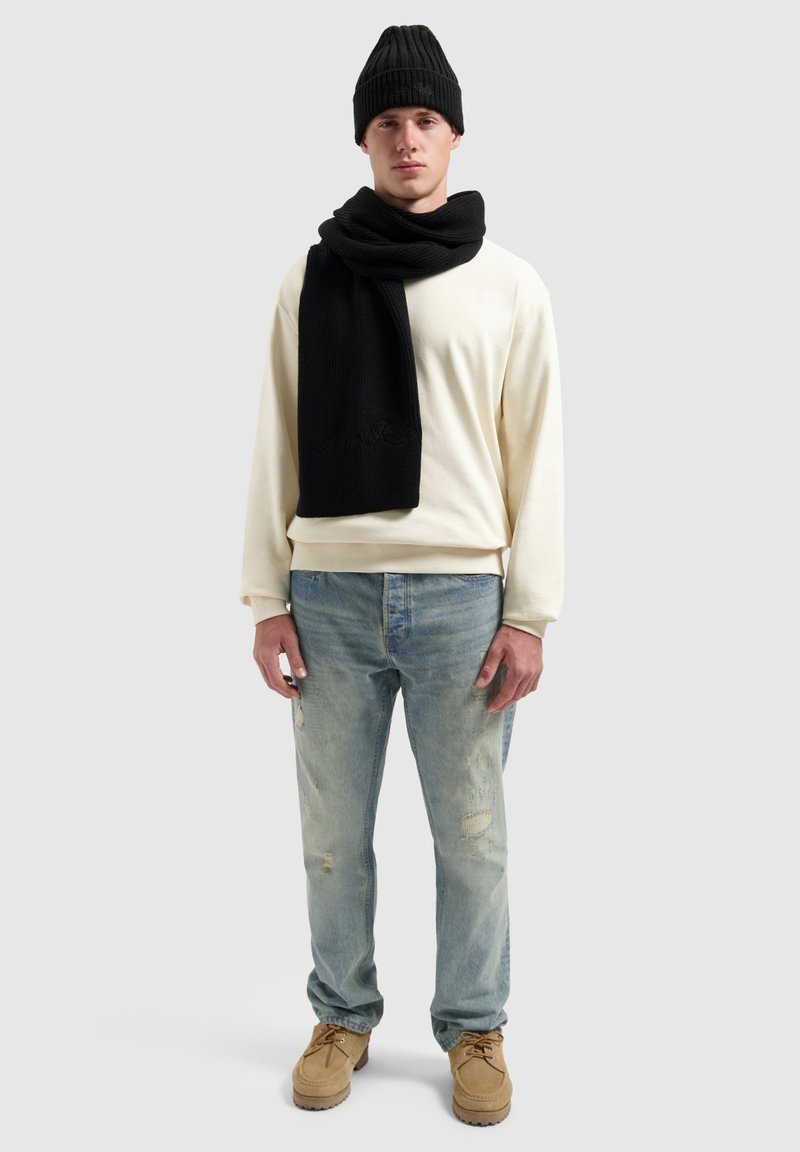Crèmekleurige sweatshirt, zwarte geribbelde breipet en sjaal, lichtblauwe distressed jeans, tan veterschoenen, staand tegen een grijze achtergrond.