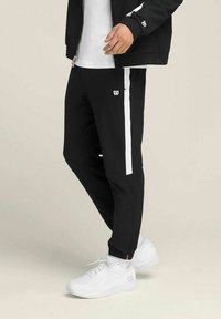 Zwarte sportieve broek met witte zijkanten, elastische boorden en een Wilson-logo. Draagt witte sneakers op een effen achtergrond.