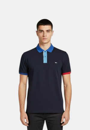Giovane uomo con indosso una polo blu navy con colletto blu, patta con bottoni azzurri, polsino della manica sinistra blu e polsino della manica destra rosso, in piedi.