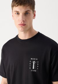 Must must have valge graafika trükkidega tuletorn ja tekst "MAKIA ESTD 2001 TTRS" vasakul rinnal.