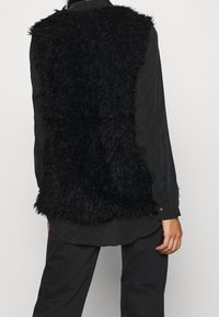 Black faux fur vest with a textured surface, worn over a dark shirt. The design is sleeveless, featuring a soft, voluminous appearance.