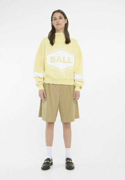 Gul sweatshirt med "BALL" grafik, cremefarvede accenter, parret med knælange beige shorts og sorte loafers. Hvide sokker synlige.