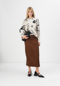 Donna che indossa una blouse floreale color crema, una gonna midi marrone, tacchi neri e tiene in mano una piccola borsa nera su uno sfondo bianco puro.