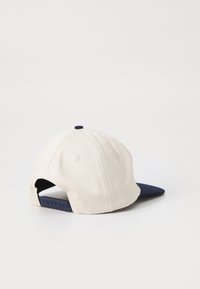 Beige színű pamut sapka sötétkék csúccsal, állítható sötétkék snapback záródással és egy kis sötétkék gombbal a tetején. Egyszerű, rendezett design.
