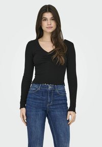 Top nero a maniche lunghe con scollatura a V e dettaglio arricciato al centro, abbinato a jeans in denim blu a vita alta. Tessuto testurizzato con bordi ondulati.
