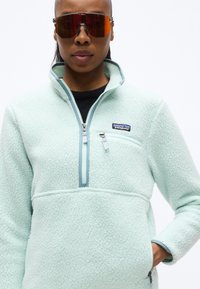 Άτομο που φοράει ανοιχτό μπλε fleece Patagonia με φερμουάρ μέχρι τη μέση και αθλητικά γυαλιά ηλίου με κόκκινο φακό, στέκεται μπροστά σε λευκό φόντο.