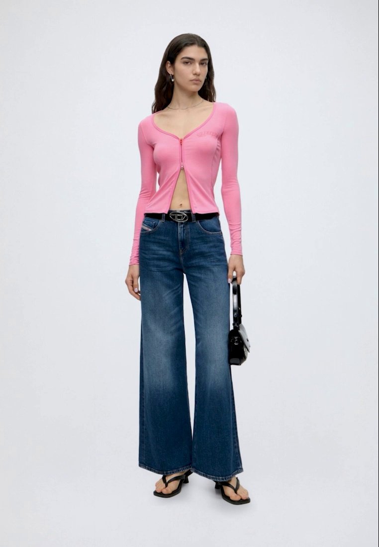 Femme debout en jean bleu évasé, haut rose boutonné et sandales, tenant un petit sac à main noir sur un fond uni.