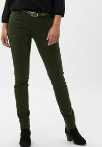 Jeans slim fit verde oliva con una textura suave, diseño de cinco bolsillos y trabillas metálicas para el cinturón. Combinados con una blusa negra y botas de tacón.