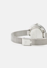 Reloj de acero inoxidable plateado con pulsera de malla, caja redonda y un cierre que presenta un logo grabado de 'CK' en el lateral.
