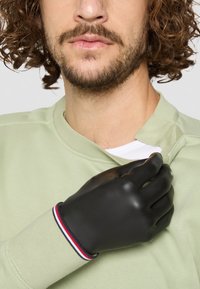 Svart latexhanske med en tilpasset design, iført på en hånd. Ved håndleddet har den et flerfarget stripete akzent mot en lysegrønn sweatshirt.