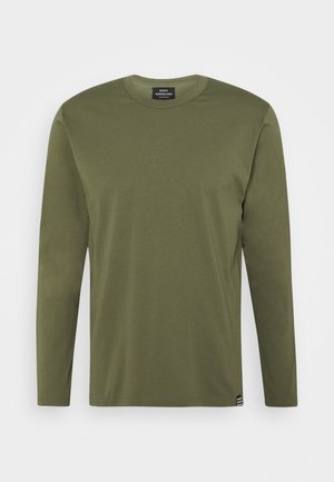 Oliivinvihreä pitkähihaisen pyöreäkauluksen paita, jossa on pieni musta-valkoinen logotagi alahelmassa valkoista taustaa vasten.